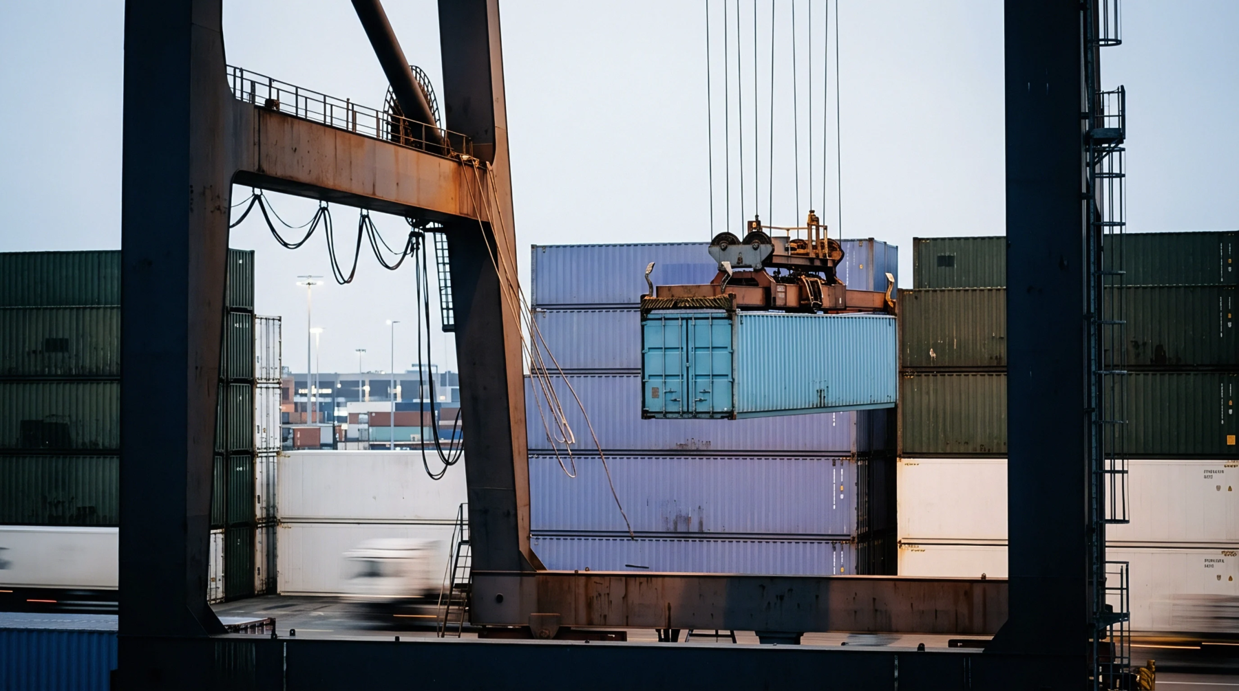 An overhead gantry crane lifts a blue shipping container into position amidst numerous stacked containers at a busy port facility.