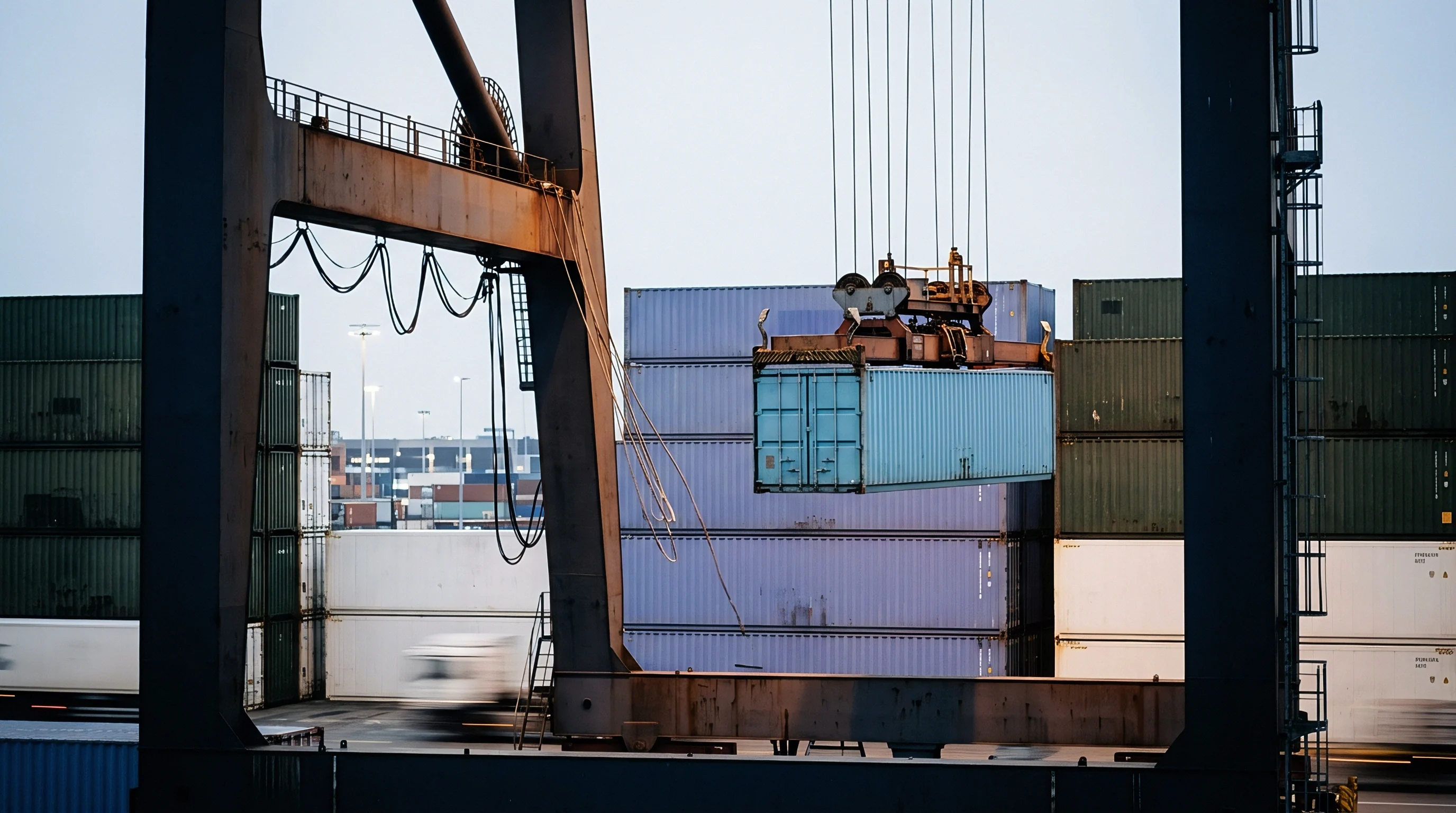 An overhead gantry crane lifts a blue shipping container into position amidst numerous stacked containers at a busy port facility.